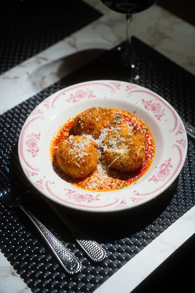 Three arancini in sunday sauce, dusted with parmigiano, on a rose-patterned plate